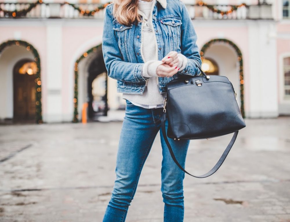 Person in a denim jacket, cream sweater and jeans holding a black leather handbag, standing on a cobblestone street