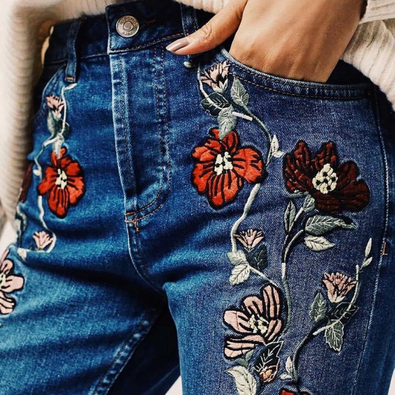 Close-up of blue denim jeans with red and pink floral embroidery, hand resting in pocket, warm knit sweater visible