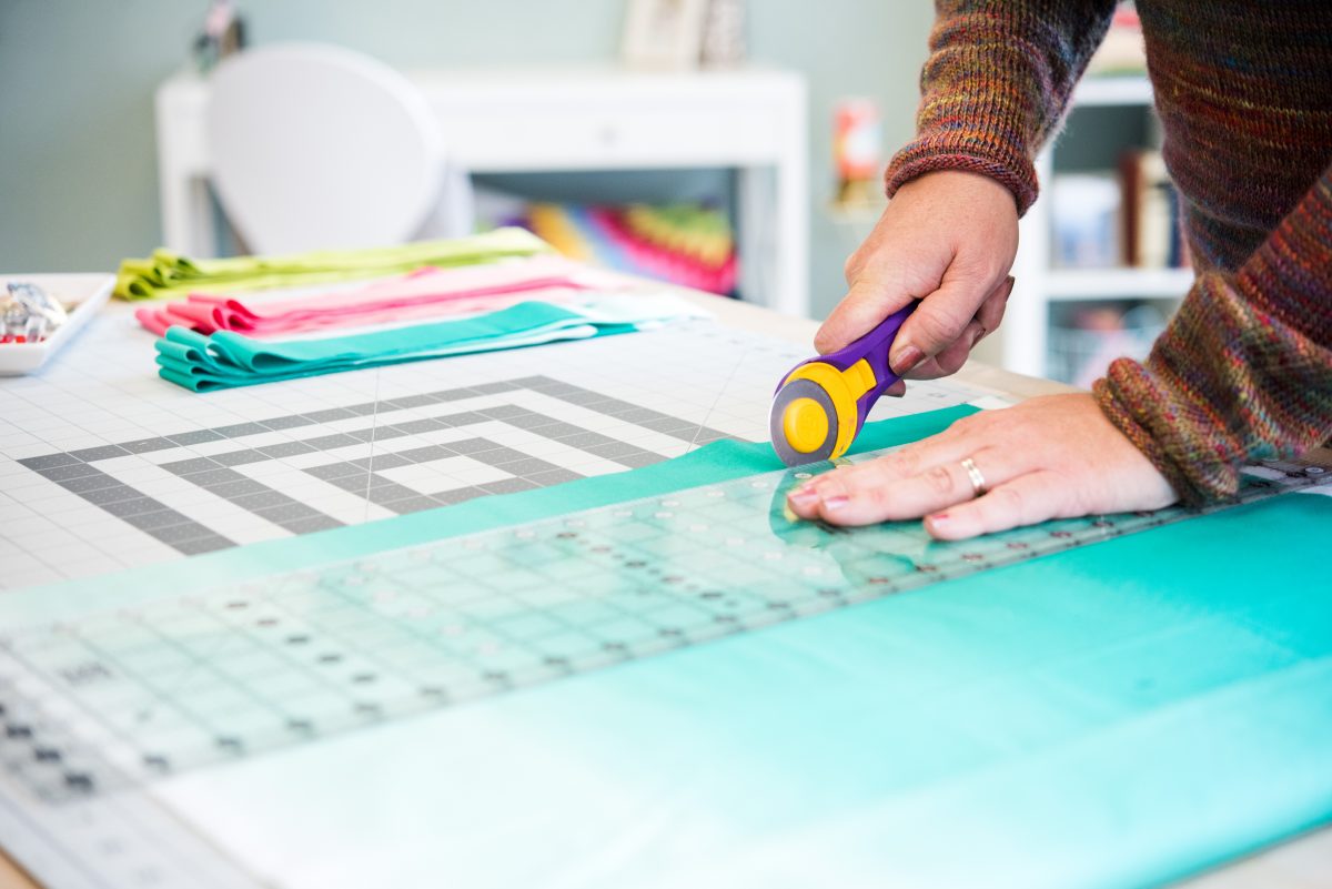 Close-up of hands using a rotary cutter to slice teal fabric on a cutting mat, with folded colorful fabrics in the background, warm and crafty mood